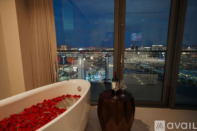 A bathtub filled with red roses sits next to a window overlooking a city at night.