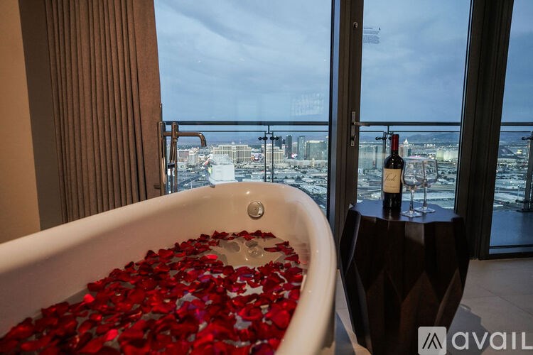 A bathtub filled with red rose petals sits in front of a window with a view of the city.