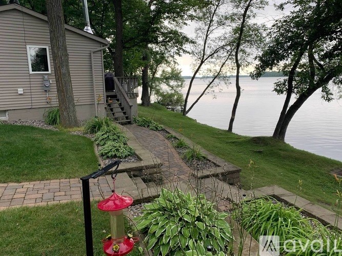 A bird feeder hangs from a pole in a garden with a house and a lake in the background.