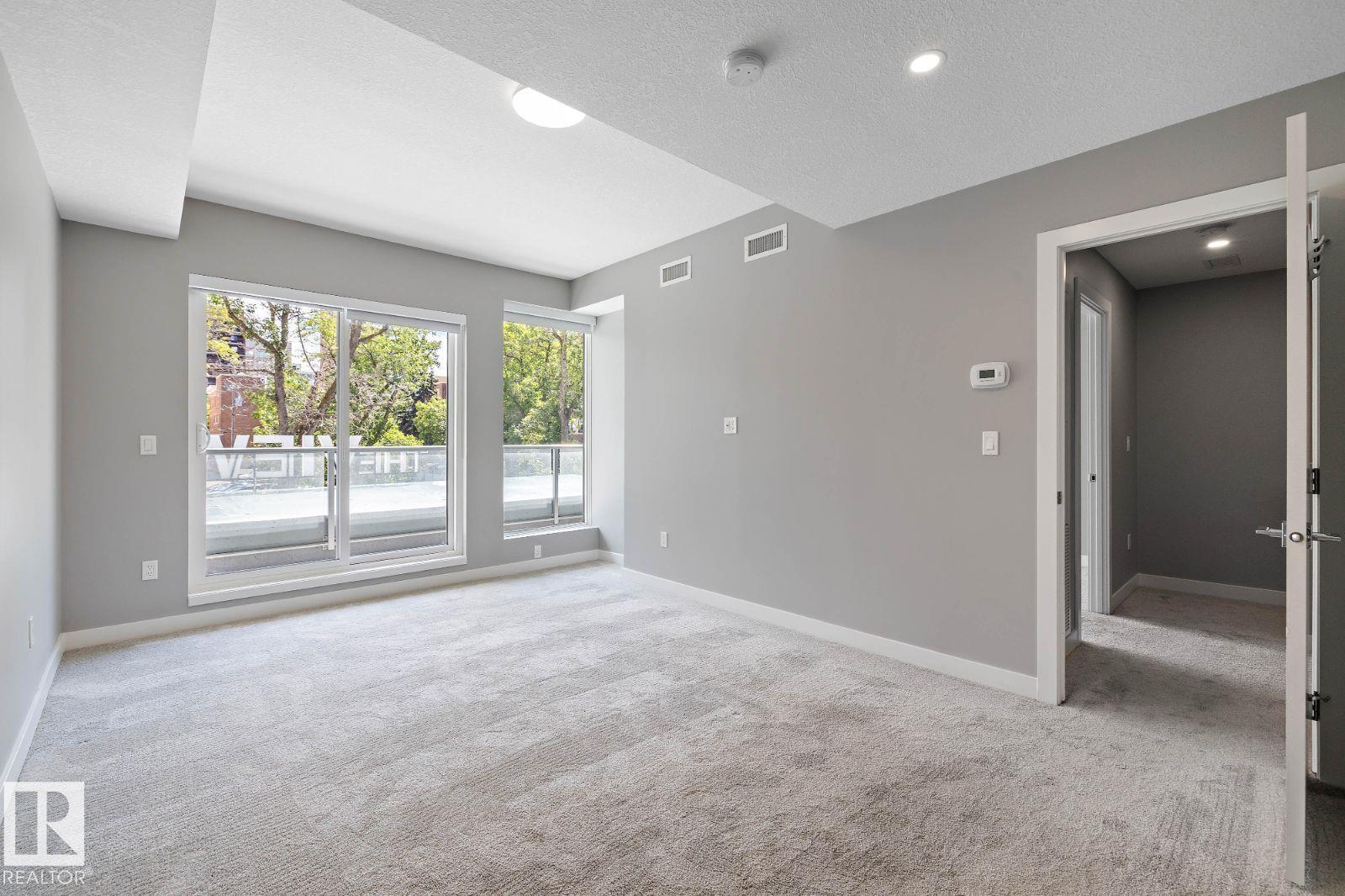 A spacious room with grey carpeting and a view of the street through the windows.