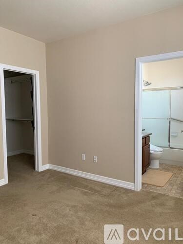 A room with beige walls and carpeted floor with a white door and a bathroom visible through the door.