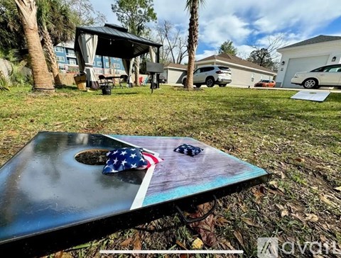 A flag-themed table with a small American flag and a black object on it is in the foreground of a yard with a house and vehicles in the background.