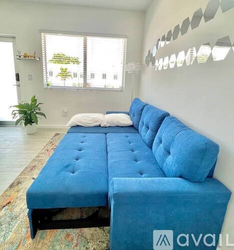 A blue couch with a white pillow is in the foreground of a room with a window and a plant.