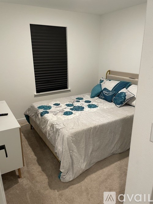 A bed with a white comforter and blue pillows is in a room with a window covered by black blinds.
