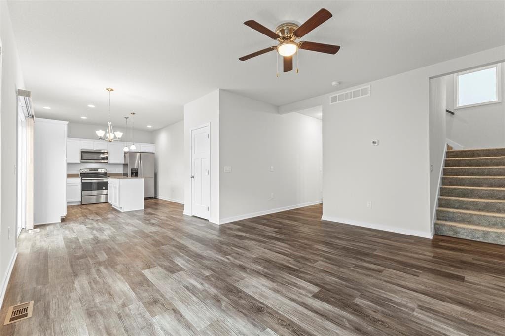 A spacious room with a ceiling fan and light fixture, hardwood flooring, and a staircase on the right side.