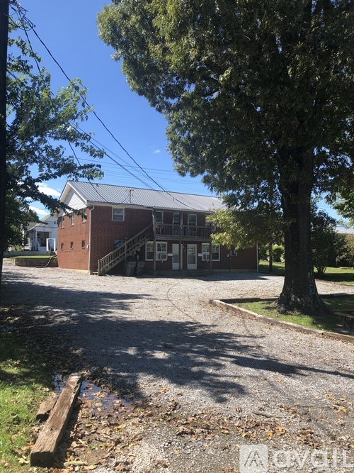 A building with a red brick exterior and a grey roof is surrounded by trees and a gravel driveway.