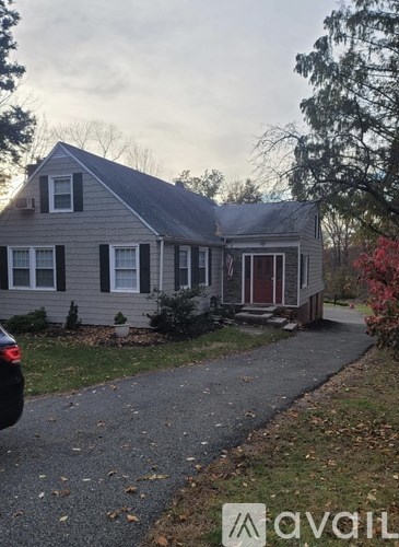 A house with a grey siding and a red door is surrounded by trees and has a driveway leading to the front door.