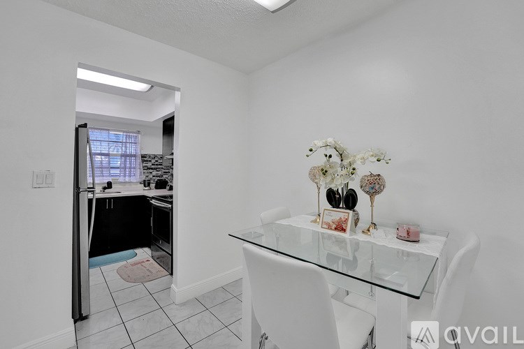 A white dining table with a glass top and white chairs in a room with a kitchen in the background.