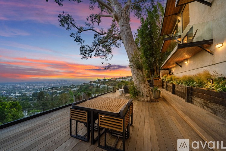 A wooden table is on a deck with a tree in front of a house.