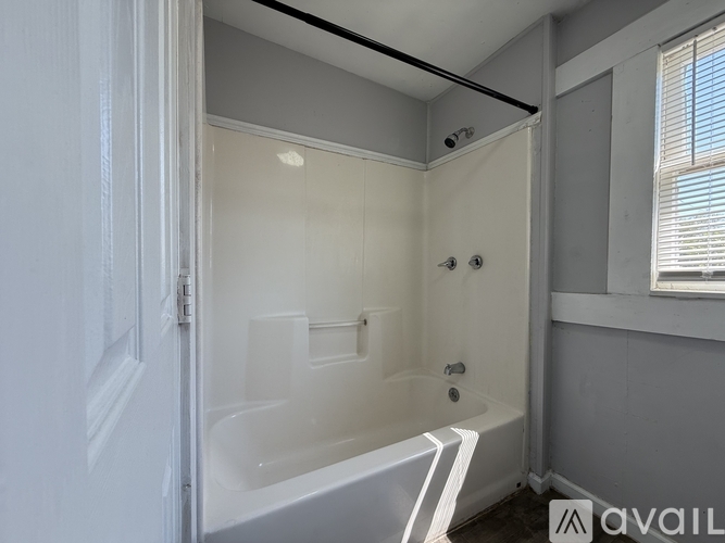A bathroom with a white tub and shower, a window with blinds, and a door.