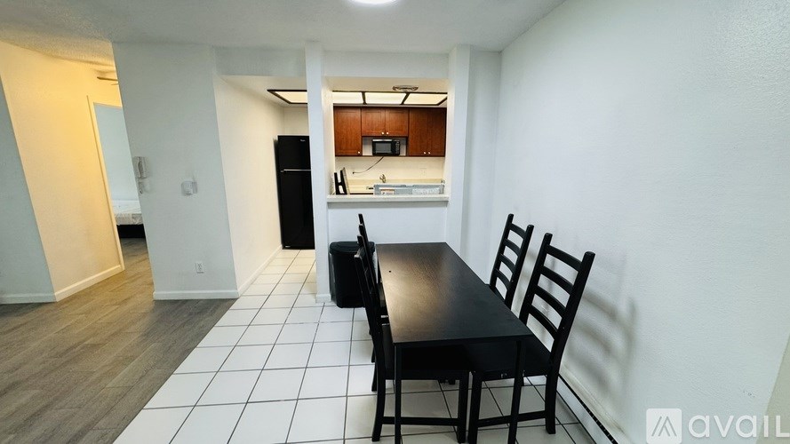 A black dining table with two chairs is in a room with white walls and a kitchen in the background.