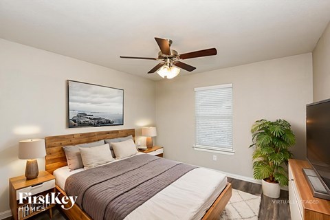 A bedroom with a bed, a ceiling fan, a nightstand with a lamp, a window with blinds, and a plant.