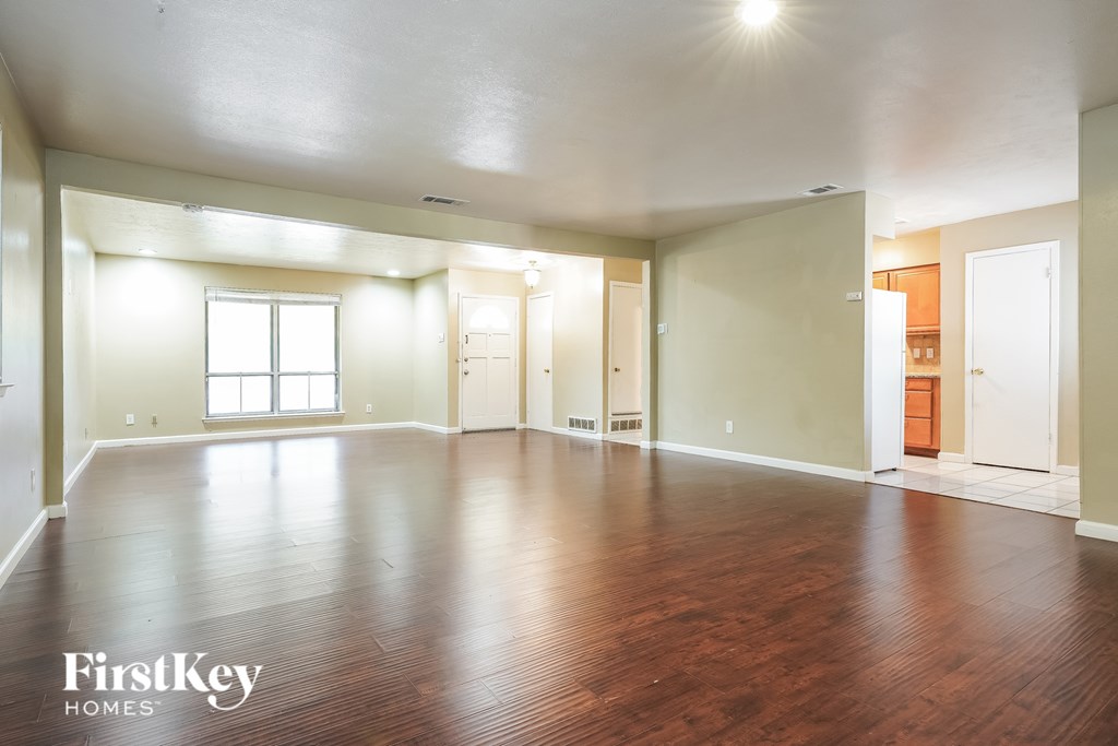A spacious room with wooden floors and white walls, with a window on the left wall.