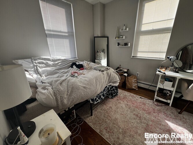 A messy bedroom with a bed covered in blankets and clothes, a desk with a computer, and a window with blinds.