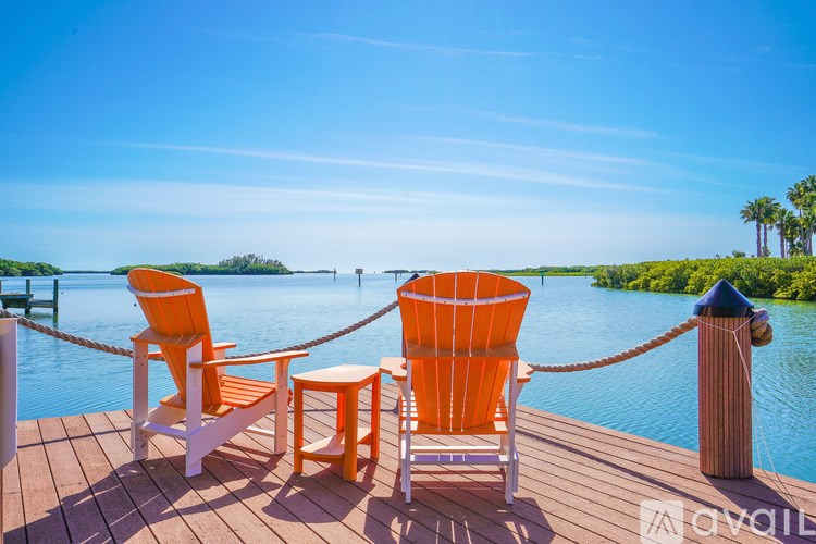 Two orange chairs and a table are on a wooden deck by the water.