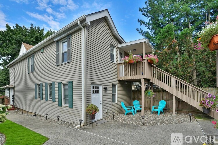 A house with a grey exterior and a white door is surrounded by a gravel driveway and a deck with chairs.