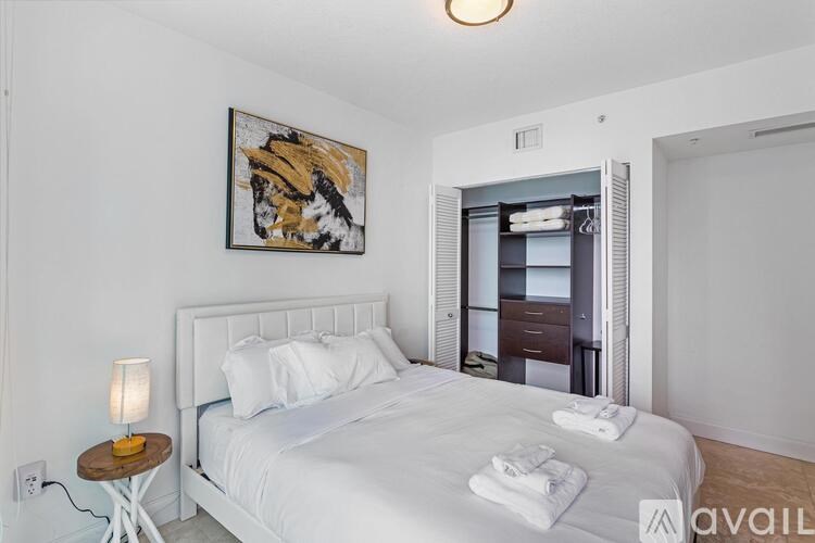 A bedroom with a white bed, a small table with a lamp, and a painting on the wall.