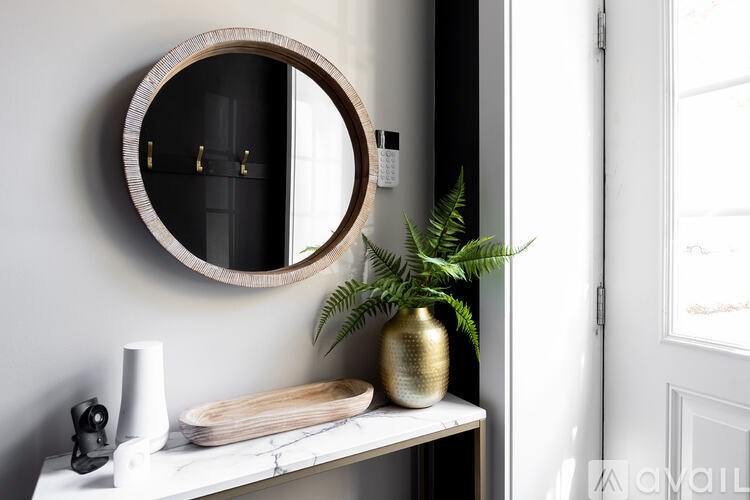 A round mirror hangs on a wall above a white shelf with a plant, a vase, and a wooden tray.