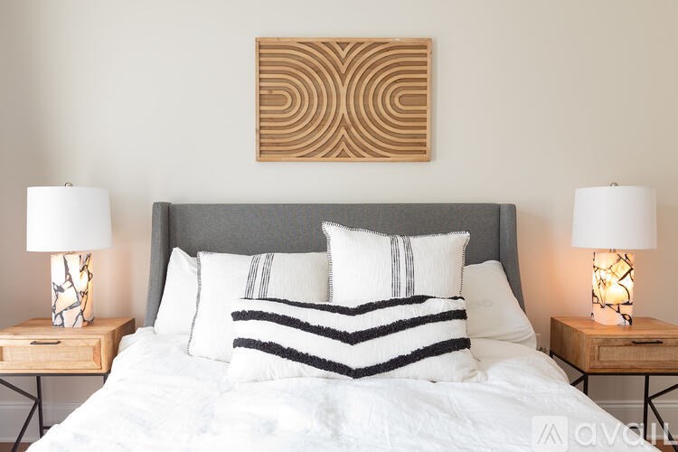 A bed with a white comforter and striped pillows is flanked by two lamps on wooden nightstands.