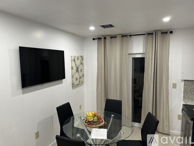 A modern dining room with a glass table and chairs, a flat screen TV, and a window with curtains.