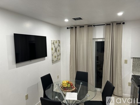 A modern dining room with a glass table and chairs, a flat screen TV, and a window with curtains.