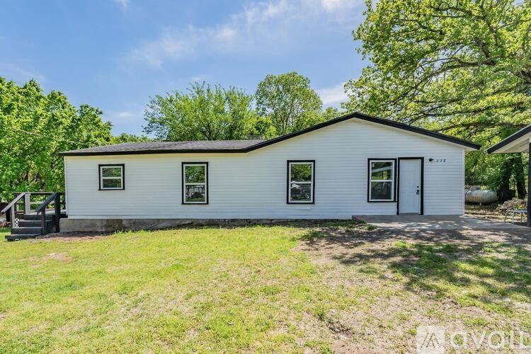 A white house with a black roof and a black door is surrounded by greenery.