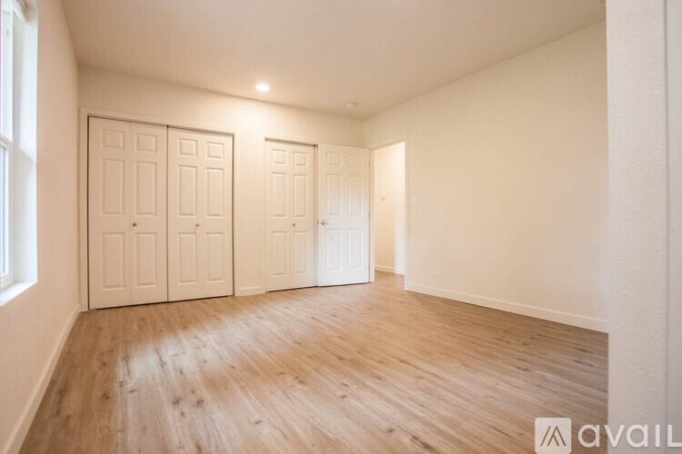 A room with wooden flooring and white walls, with a window on the left and three doors on the right.
