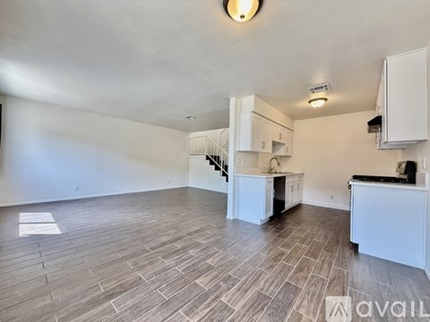 A spacious room with wooden flooring and white walls, featuring a kitchenette with white cabinets and appliances.
