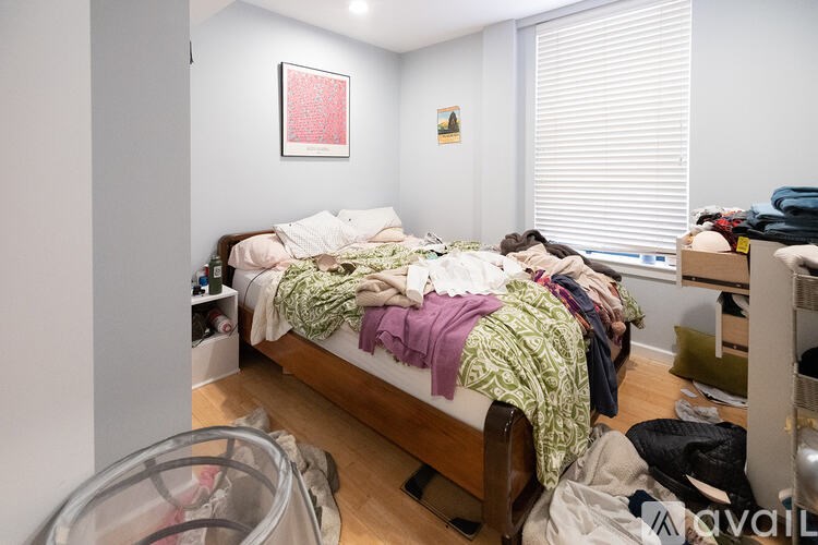 A messy bedroom with a bed covered in clothes and a window with blinds.