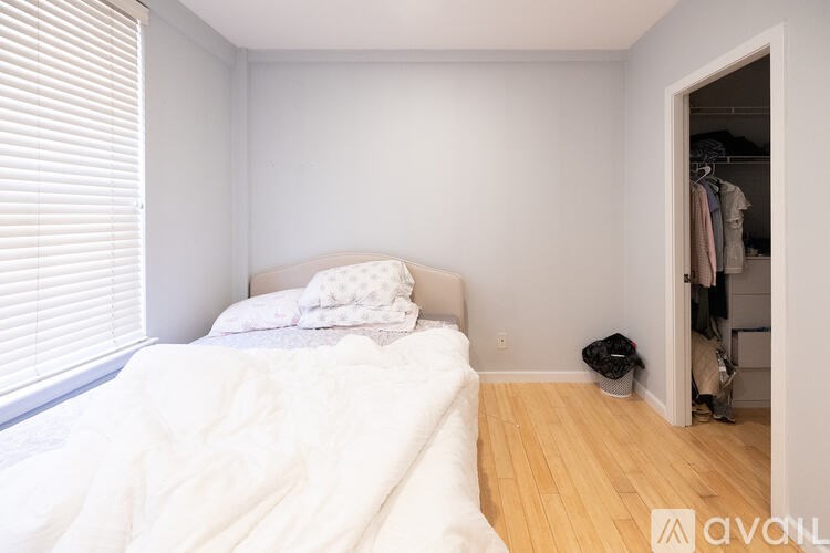 A bedroom with a bed, a window with blinds, a wooden floor, and a closet with clothes.