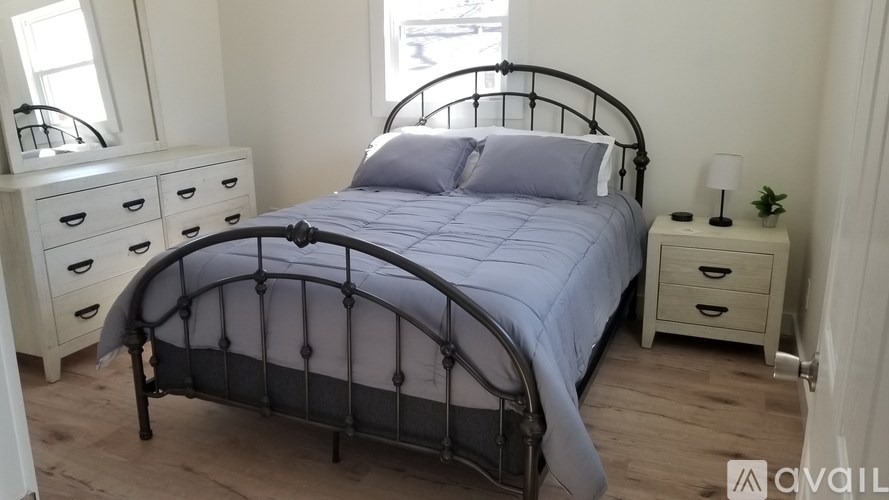 A bed with a metal frame and a grey bedspread is in the foreground of a room with a white dresser and a small plant on a side table.