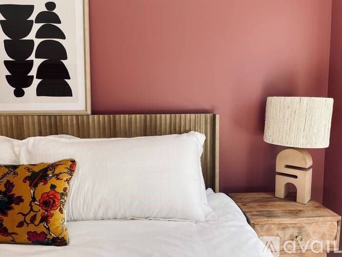 A bed with a white pillow and a floral pillow is in front of a red wall with a framed picture of black shapes above it.