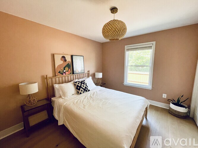 A bedroom with a large bed, a nightstand with a lamp, a window, and a plant.