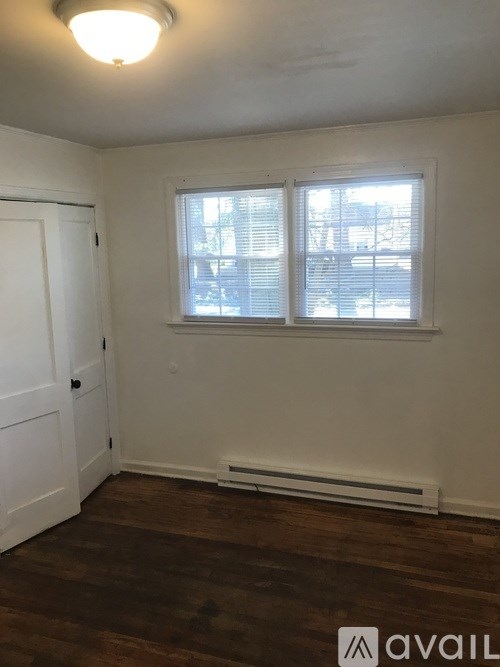 A room with a white door, a window with blinds, and a wooden floor.