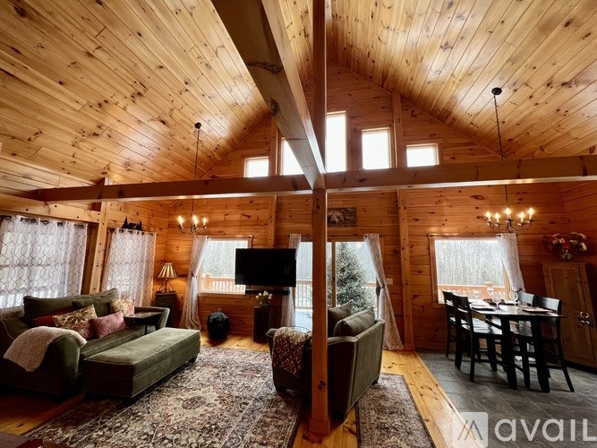 A cozy living room with wooden walls and ceiling, furnished with a sofa, armchair, and a dining table set for four.