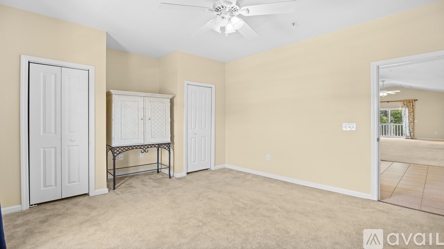 A room with beige walls and carpeting, featuring a ceiling fan and a table with chairs.