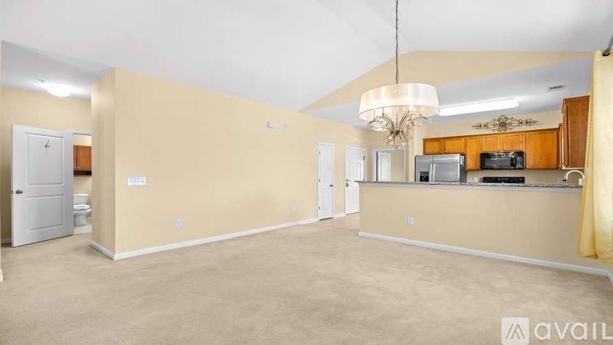 A spacious living room with beige walls and carpeting, featuring a kitchen area with wooden cabinets and stainless steel appliances.