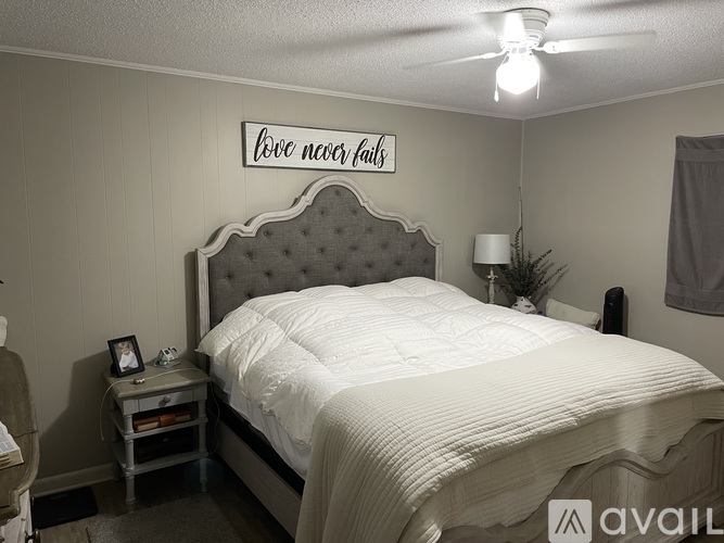 A bedroom with a bed, a nightstand with a picture frame, and a wall decoration that says "love never fails".