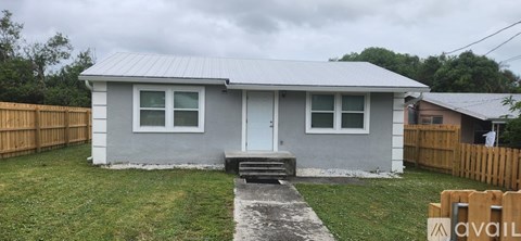 A small house with a grey roof and a white door is surrounded by a wooden fence.