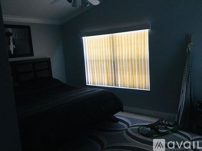A room with a bed, a window with blinds, a ceiling fan, and a rug with a shoe on it.