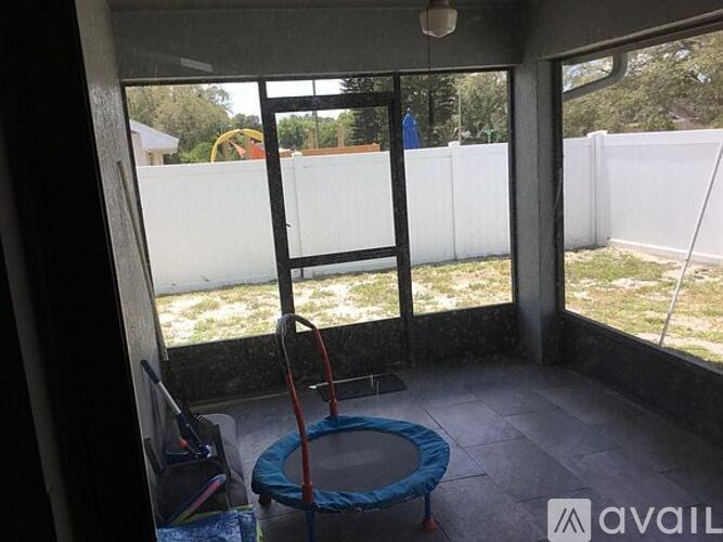 A blue and red swing set sits in a sunny room with a white fence and trees visible through the window.