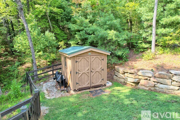 A wooden outhouse with a green roof is surrounded by a stone wall and trees.