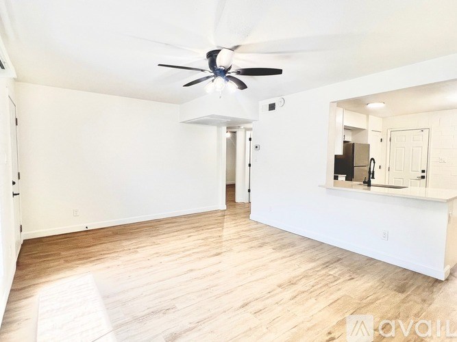 A spacious room with a ceiling fan and light, hardwood floors, and a kitchenette in the background.