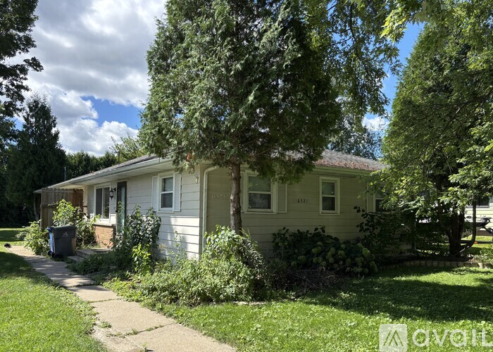 A house with a white exterior is surrounded by greenery and has a trash can in front.