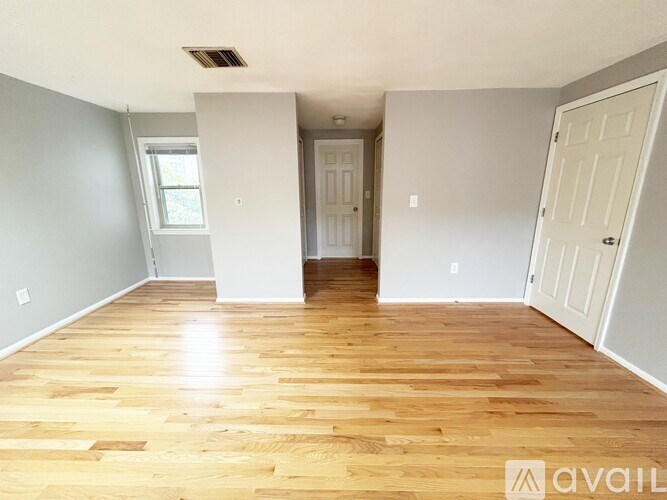 A room with wooden flooring and white walls, with a door on the right and a window on the left.