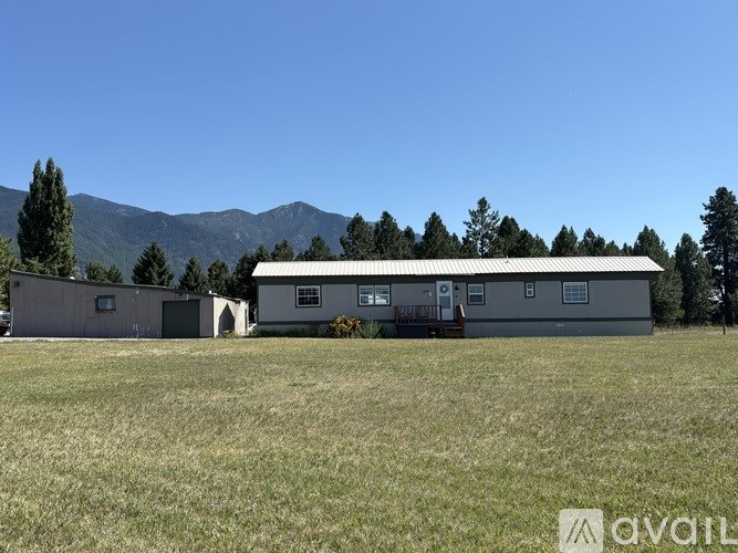 A large building with a metal roof is situated in a grassy field with trees and mountains in the background.