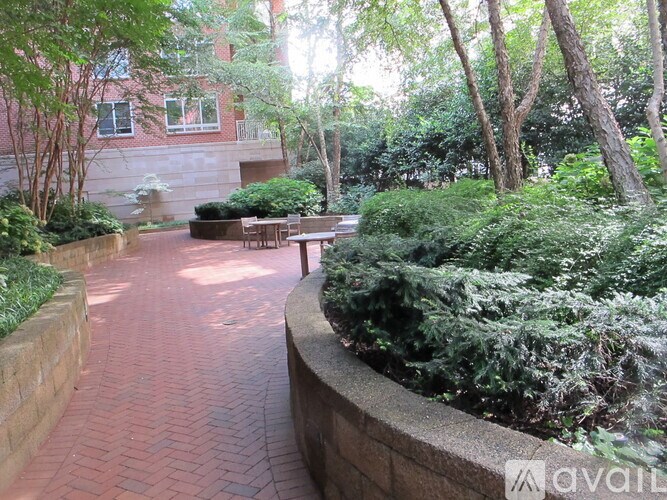 A brick walkway leads through a landscaped area with a bench and a building in the background.