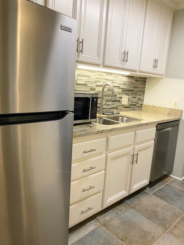 A stainless steel refrigerator stands in a kitchen with white cabinets and a black dishwasher.