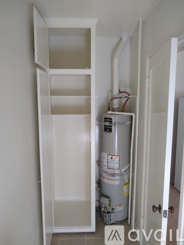 A white door is open to a room with a white wall and a white shelving unit with a silver water heater under it.