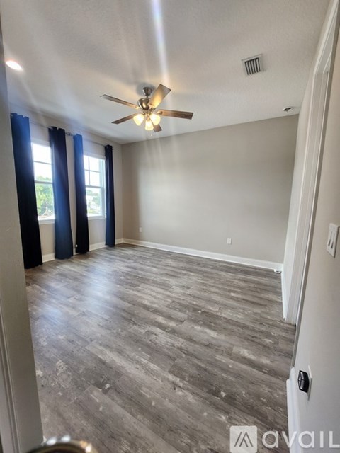 A room with a ceiling fan and light, hardwood floors, and a window with blue curtains.